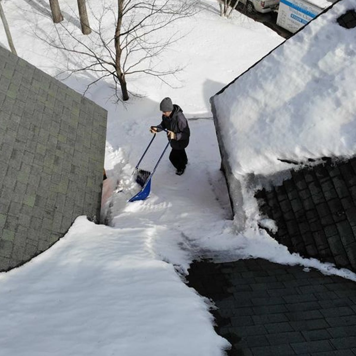 A person wearing winter clothing is shoveling snow from a rooftop walkway between two buildings, surrounded by snow-covered roofs and trees.