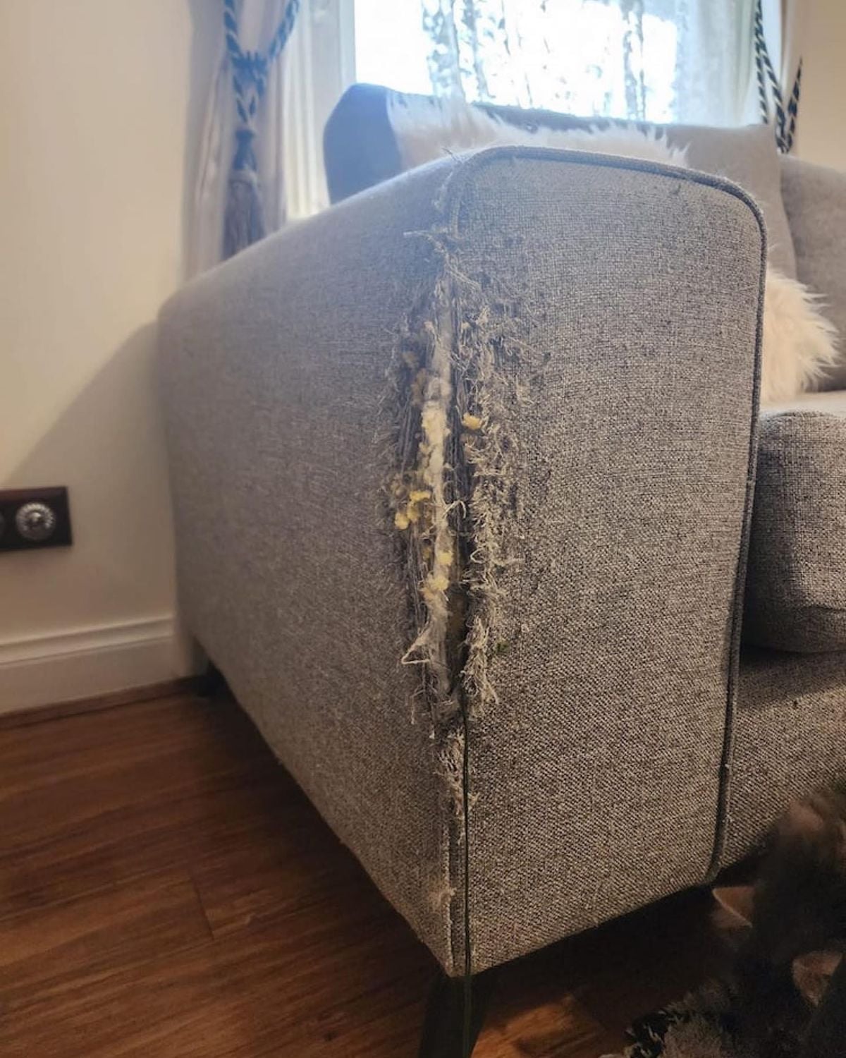 A grey fabric sofa with a severely scratched and torn armrest, exposing the foam and inner material. The damage appears to be caused by an animal, likely a cat. The sofa is in a room with wooden flooring.
