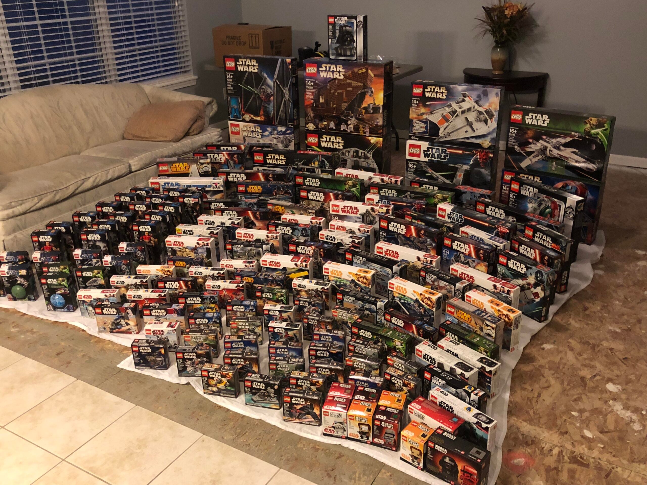 Dozens of LEGO Star Wars sets, both large and small boxes, are neatly arranged on the floor and furniture in a living room, covering a white sheet. A beige couch and a vase sit in the background.