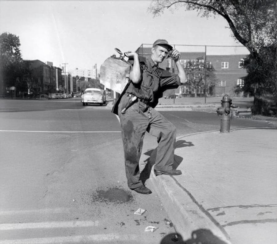 A man in work clothes stands on a curb, drinking from a bottle and carrying a large metal container over his shoulder. He is on a city street with buildings, a fire hydrant, and cars in the background.