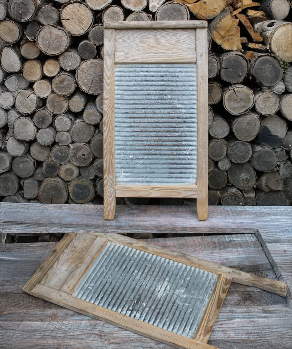 A vintage metal washboard with a wooden frame stands upright, while another similar washboard lies flat. Both are set against a background of stacked firewood logs.
