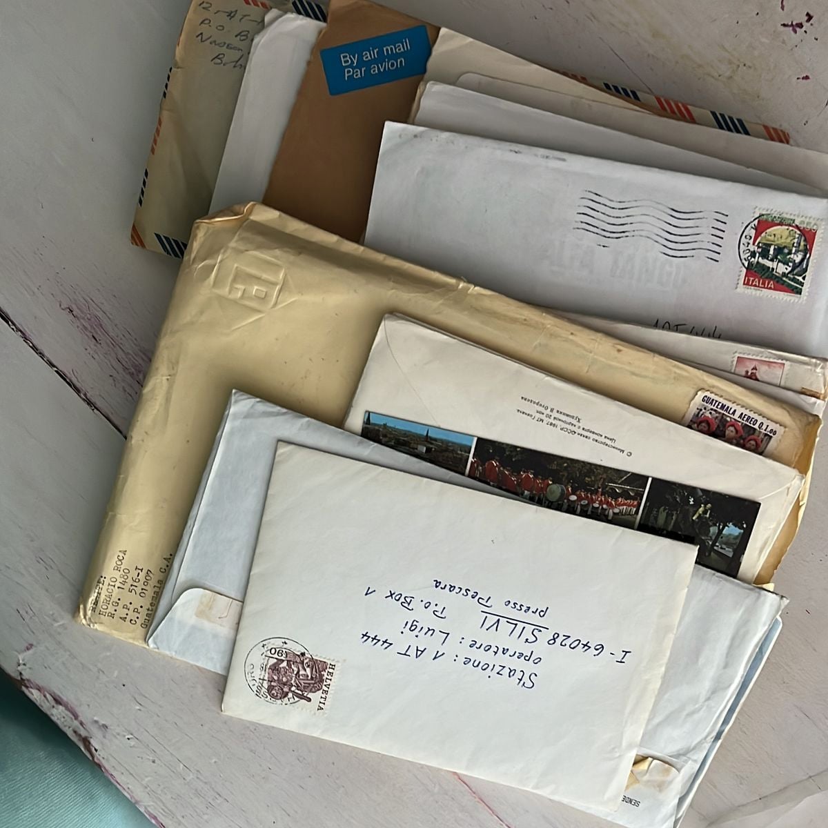 A stack of assorted envelopes and letters, including some airmail envelopes and various stamps, resting on a white wooden surface. Handwritten addresses are visible on some of the envelopes.
