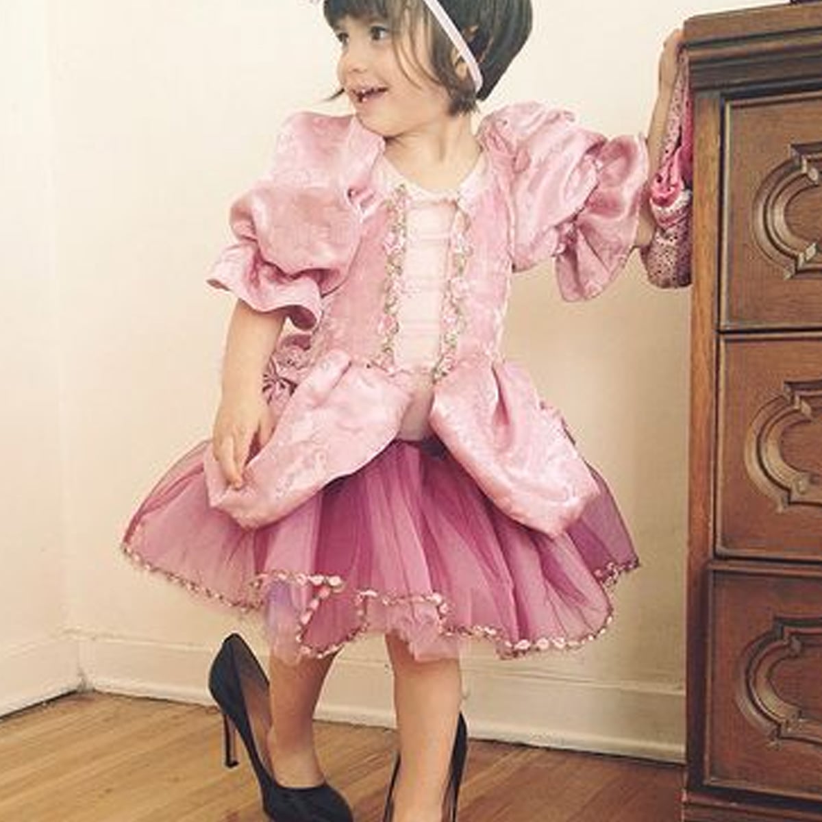 A young child wearing a pink princess dress, headband, and oversized black high heels smiles while standing near a wooden dresser in a room with light-colored walls and floor.
