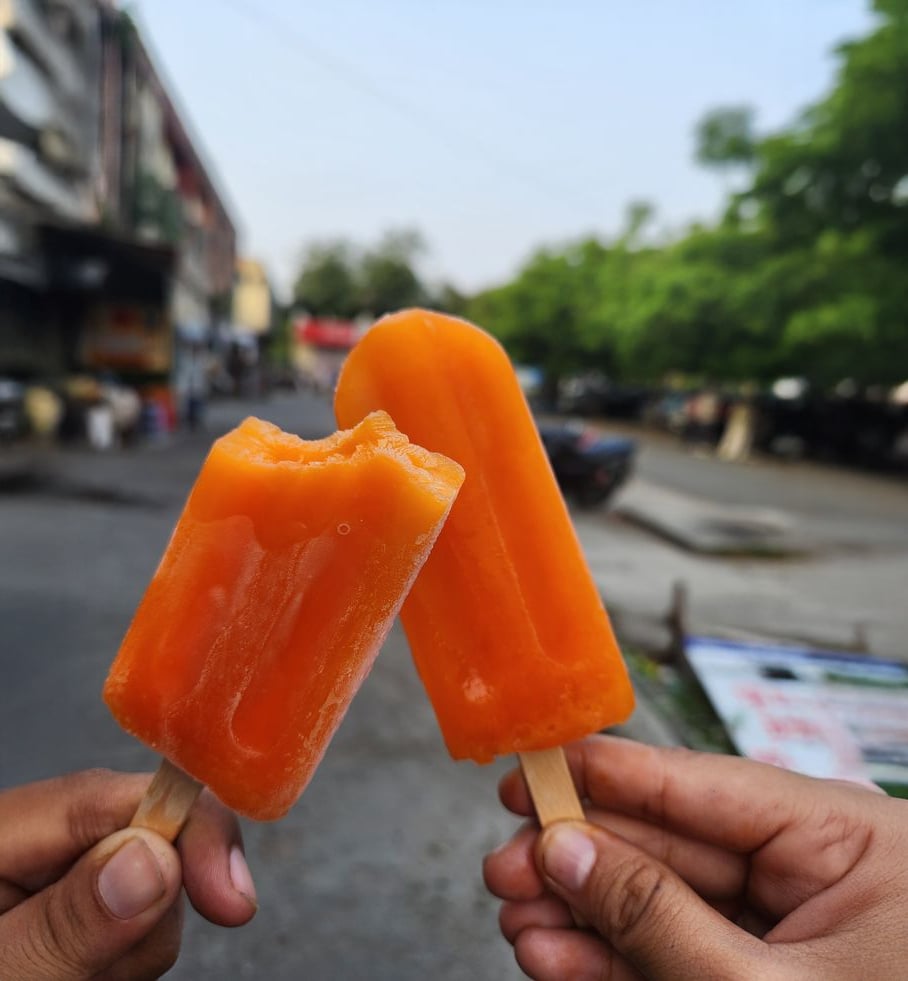 Two hands holding orange popsicles outdoors, with one popsicle showing a bite. The background features a street, trees, parked cars, and buildings, slightly out of focus.