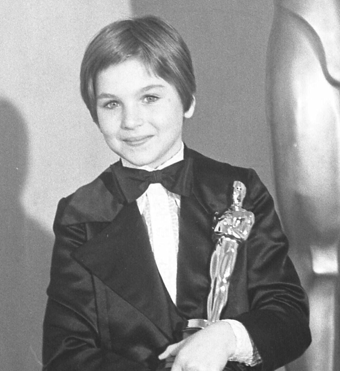 A young child wearing a tuxedo smiles while holding an Oscar statuette, standing in front of a large Oscar statue. The photo is in black and white.