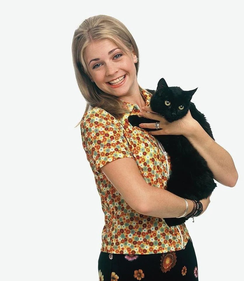 A smiling young woman with long blonde hair, wearing a floral short-sleeved shirt, holds a black cat in her arms against a plain white background.
