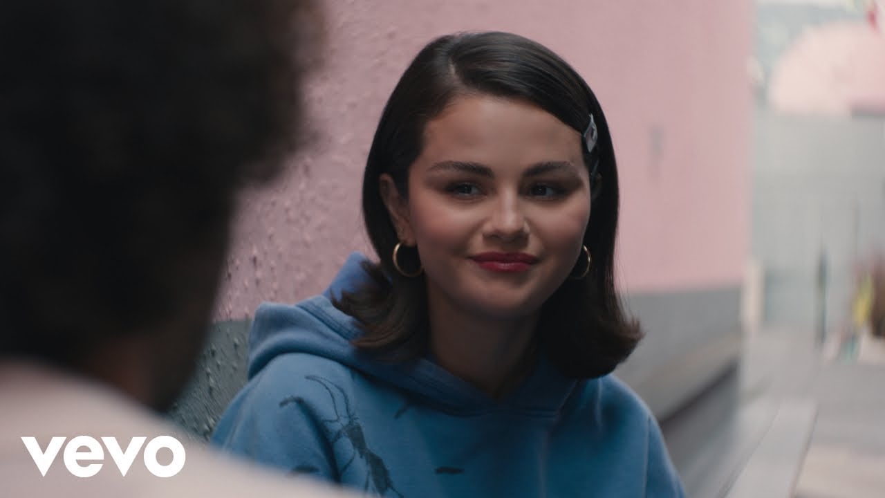 A woman with dark hair and a blue hoodie smiles slightly while sitting outdoors against a pink wall. She wears hoop earrings and a hair clip. The image has the Vevo logo in the corner.