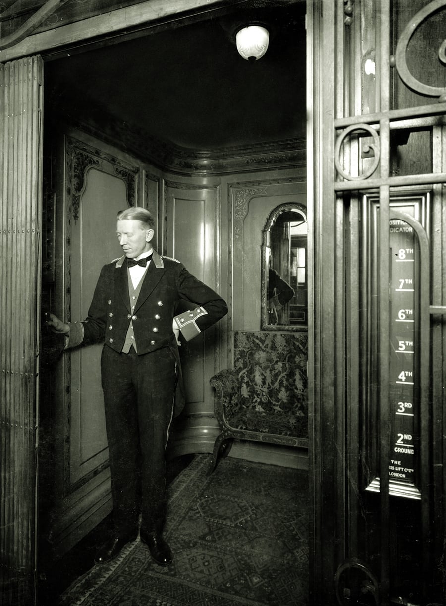 A uniformed elevator operator stands at the open door of an ornate vintage elevator, holding a lever. The elevator interior is decorated with intricate woodwork and a cushioned bench. A floor indicator is visible on the right.