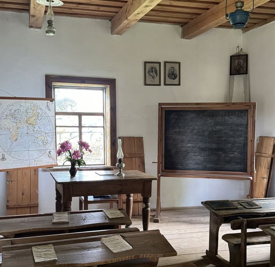 A vintage classroom with wooden desks, a chalkboard, a world map, an oil lamp on the teacher’s desk, and flowers in a vase. Sunlight comes through the window and portraits hang on the wall above the chalkboard.