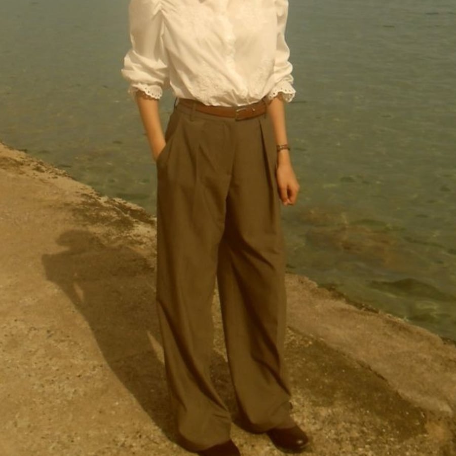 A person stands by the water on a sunlit concrete path, wearing a white long-sleeve blouse, brown wide-leg trousers, a brown belt, and black shoes. Their hands are in their pockets, and their face is not visible.