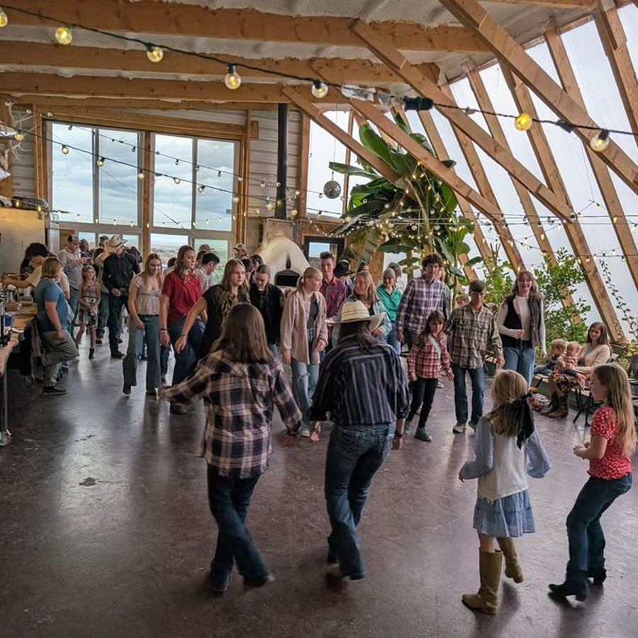 A group of people, including children and adults in casual and plaid clothing, dance together in a rustic, light-filled room with large windows and string lights, creating a lively, communal atmosphere.