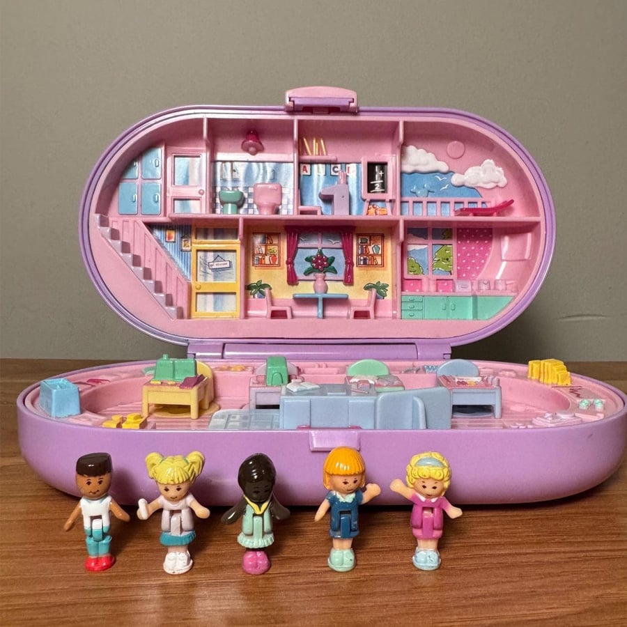 A pink Polly Pocket playset is open, revealing a detailed miniature house interior. Five small, colorful dolls stand in front of the playset on a wooden surface. The background is a plain grey wall.