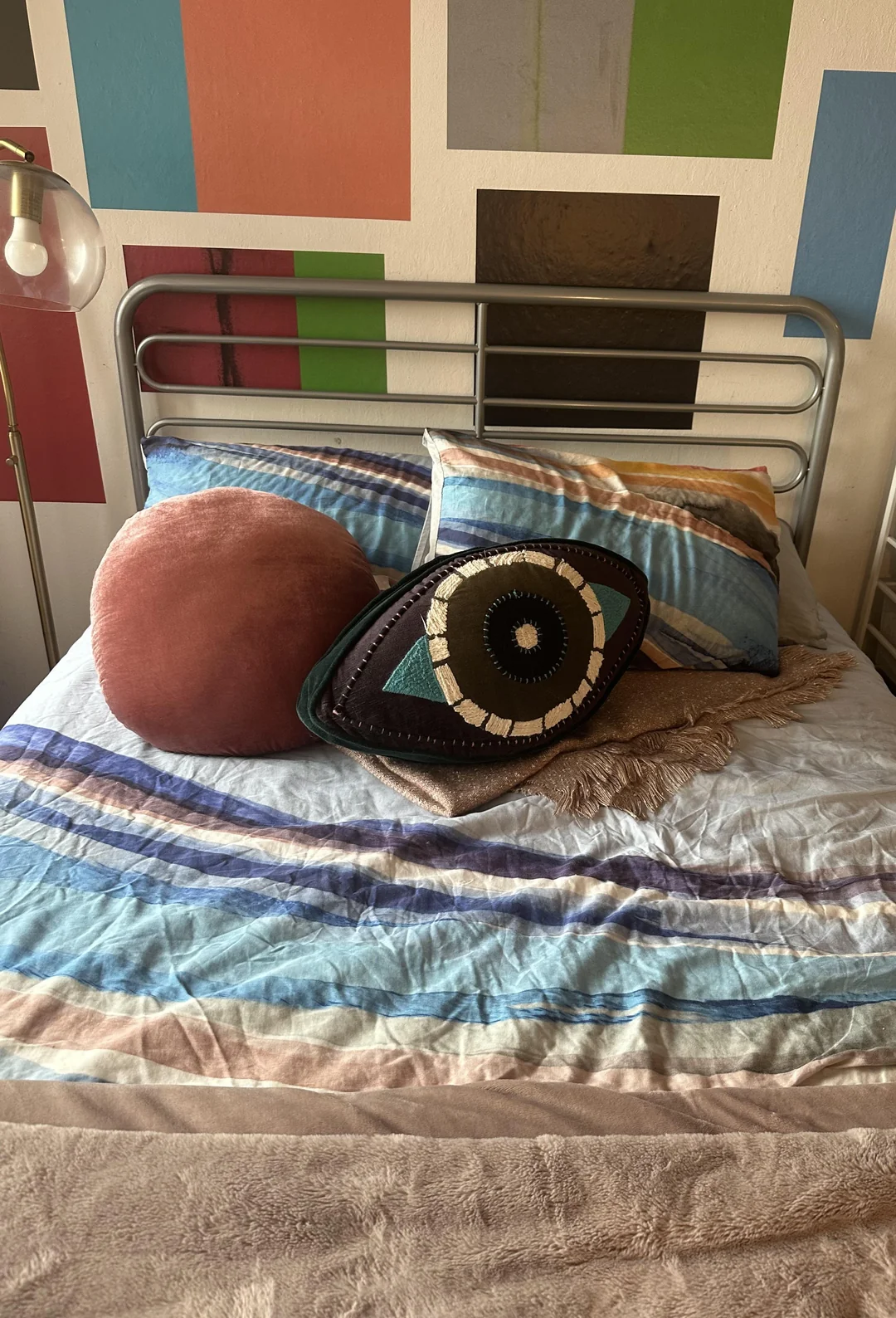 A neatly made bed with a blue and brown abstract-patterned duvet, a round pink pillow, and an eye-shaped pillow. Behind the bed is a metal headboard and a wall with colorful rectangular designs.