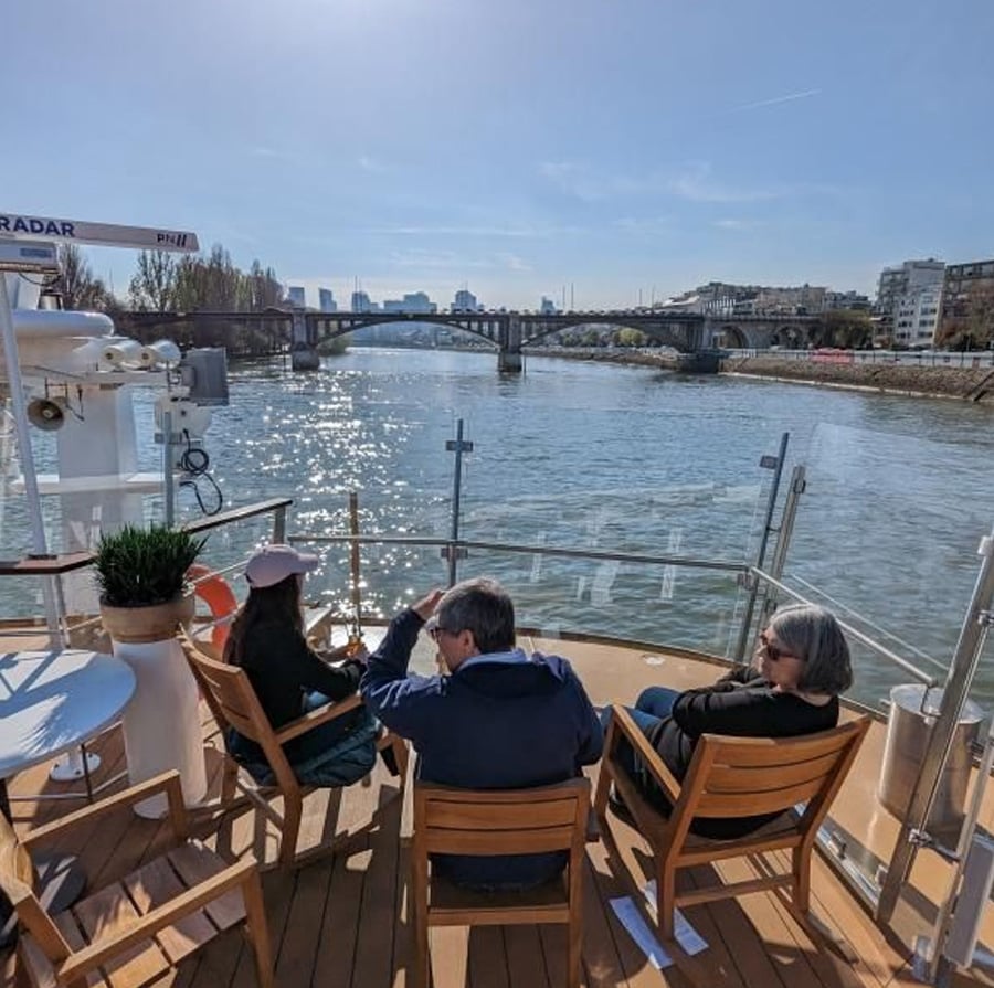 Three people sit on wooden chairs at the back of a boat cruising along a river, with a city skyline, bridges, and blue sky in the background. Sunlight reflects off the water.