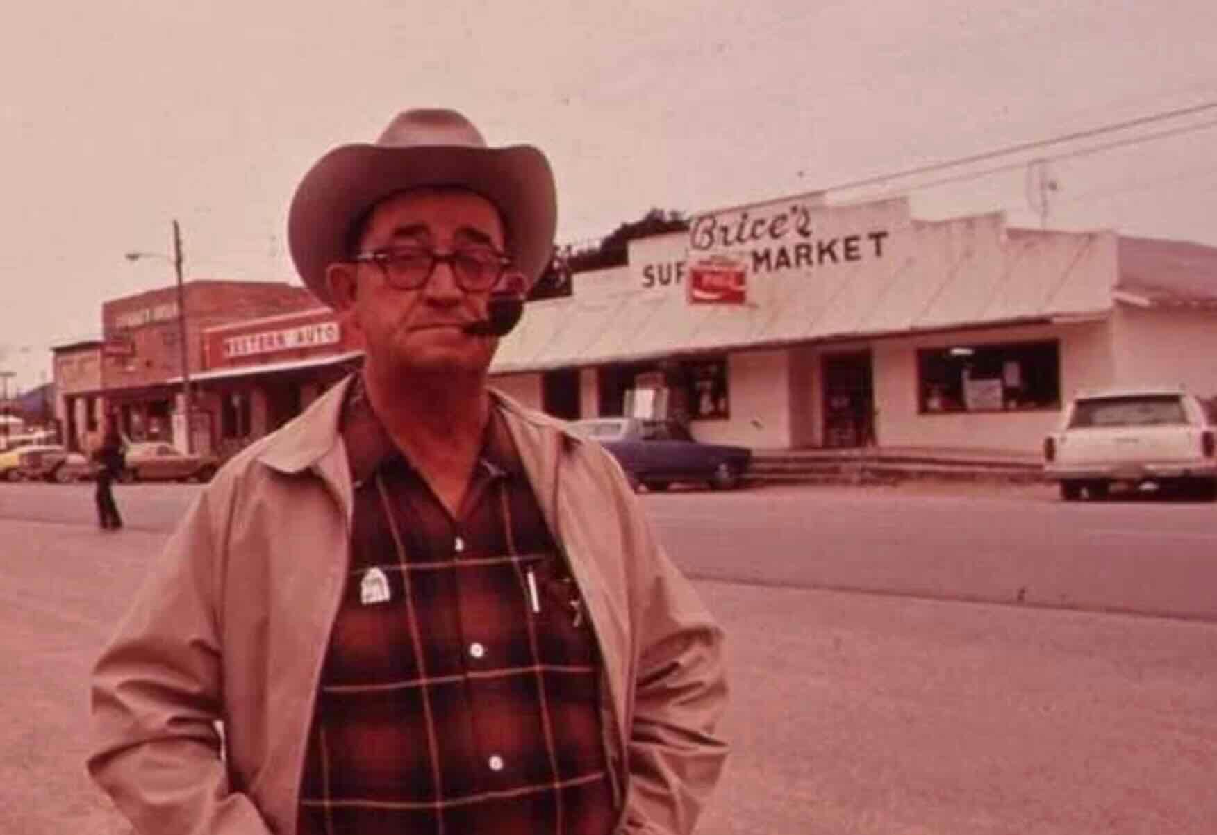 An older man in a cowboy hat, glasses, and plaid shirt stands on a street, smoking a pipe. Behind him is Brice’s Super Market and other storefronts, with cars parked along the road. The scene has a vintage feel.