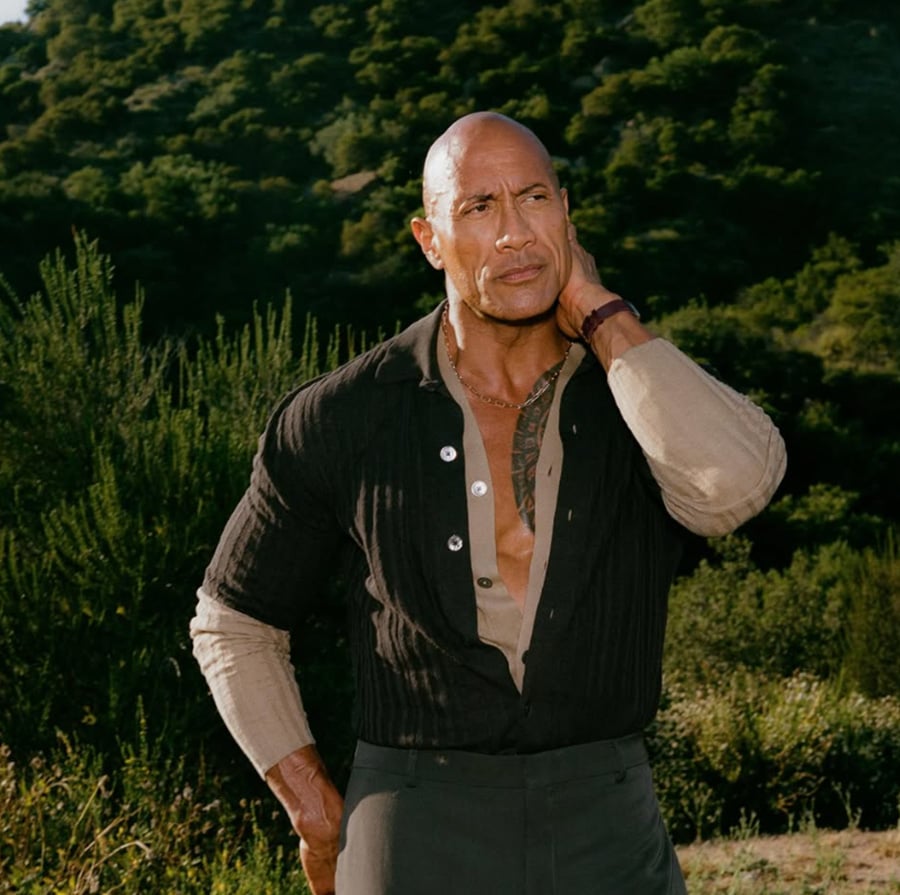 A bald man wearing a partially unbuttoned black shirt stands outdoors in front of green, tree-covered hills, with one hand touching his neck and a thoughtful expression on his face.