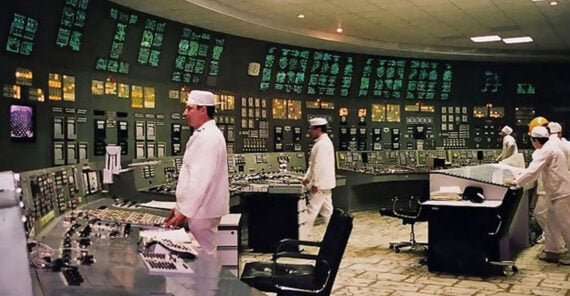 Engineers in white uniforms and hats operate a large control room filled with numerous illuminated buttons, switches, and display panels on curved walls. Some sit at desks, while others monitor instruments and controls.