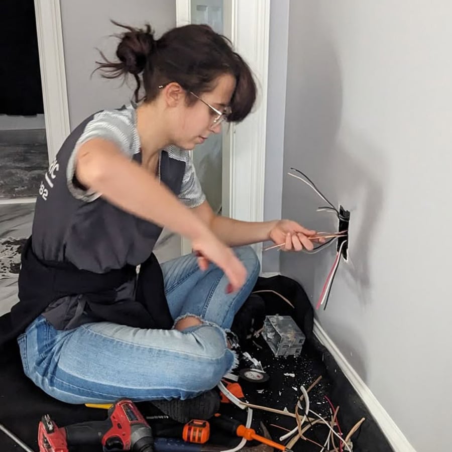A person wearing glasses and jeans is kneeling beside a wall, working on exposed electrical wires coming out of an outlet box. Tools and wiring parts are scattered on the floor nearby.