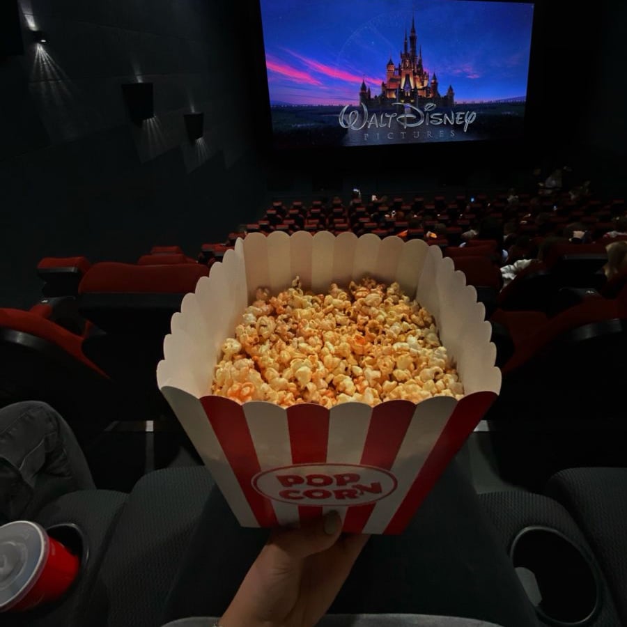 A person holds a striped popcorn box in a dark movie theater, facing a large screen showing the Walt Disney Pictures logo. A red drink cup sits beside them, and other viewers are seated ahead.