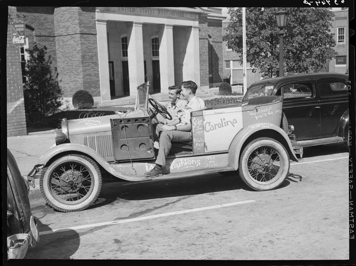 Two young men sit in an old convertible car covered in writing and drawings, parked on a sunny street in front of a U.S. Post Office building. Another car is parked nearby.