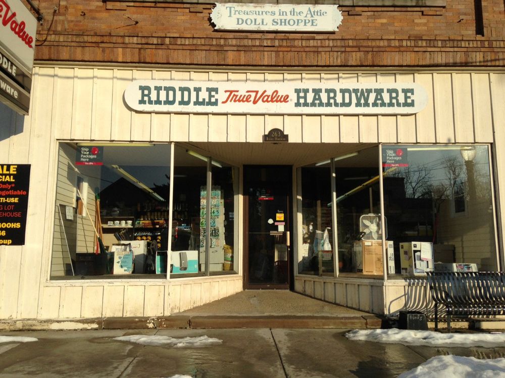 Front view of Riddle True Value Hardware store with large windows, a sign above the entrance, and a "Treasures in the Attic Doll Shoppe" sign above. Patches of melting snow are on the sidewalk outside.