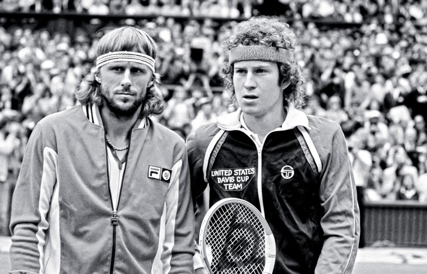 Two male tennis players in tracksuits and headbands stand side by side on a tennis court, holding rackets, with a large crowd of spectators in the background. The image is in black and white.