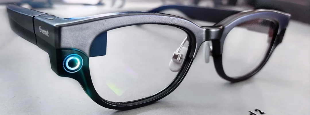 A pair of black smart glasses with thick frames sits on a table. A small circular light glows blue on the left side of the frames, indicating the device is powered on.