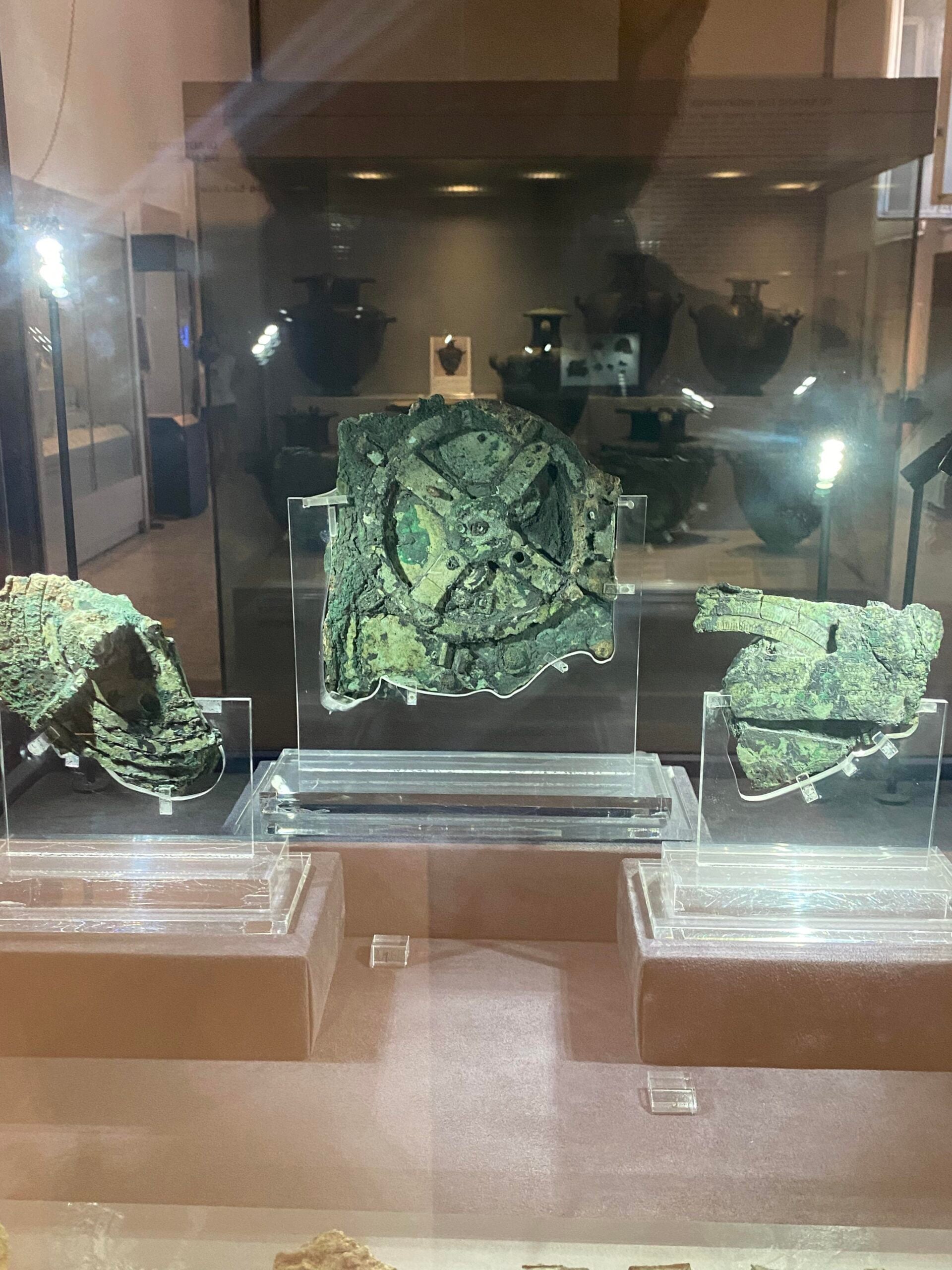 Three corroded bronze fragments of the ancient Antikythera mechanism displayed in a glass case, with the central piece showing visible gears and mechanical parts, in a museum setting.
