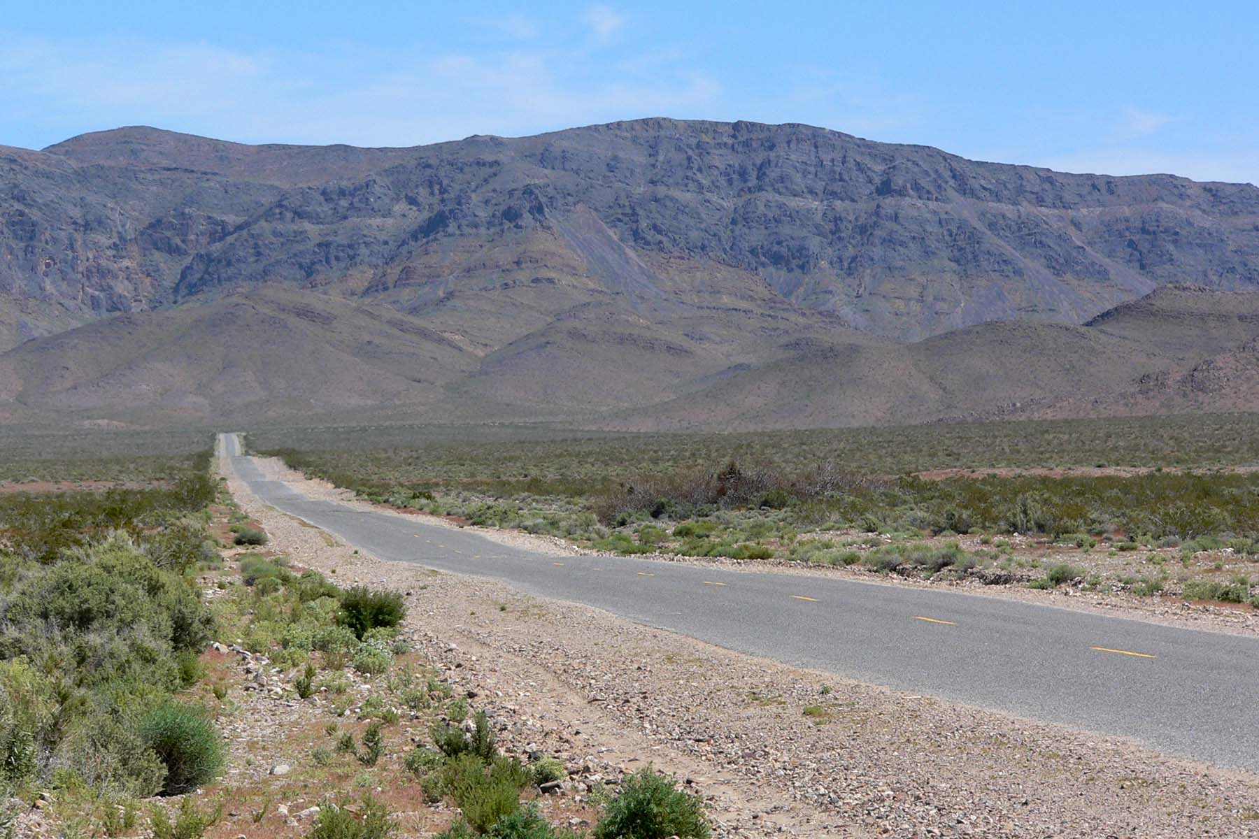 A paved, empty road stretches into the distance through a dry, rocky desert landscape with sparse vegetation and rugged mountains in the background under a partly cloudy sky.
