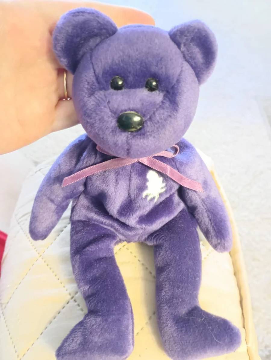A hand holds a purple plush teddy bear with black eyes, a black nose, a purple ribbon around its neck, and a white embroidered rose on its chest. The bear is sitting on a quilted white surface.
