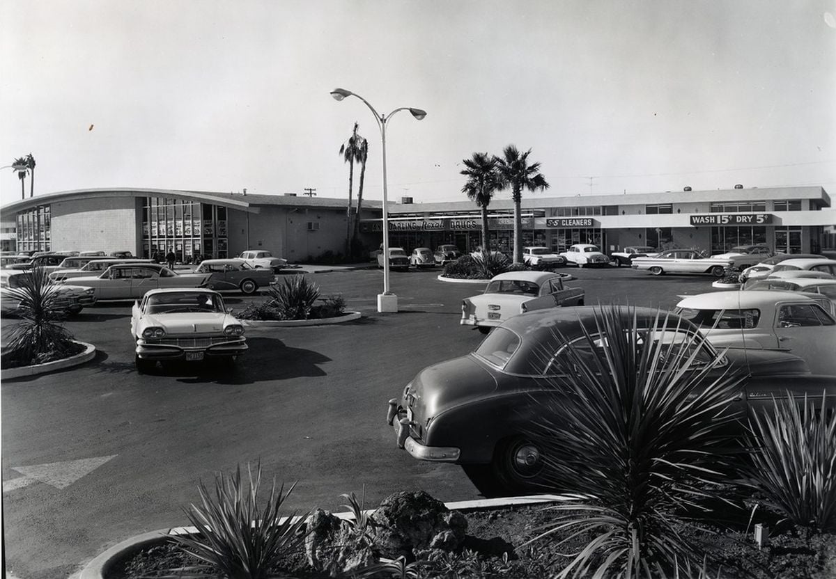 Black and white photo of a mid-20th-century shopping center parking lot, with classic cars parked and palm trees around. Store signs and a curved building are visible in the background. Desert landscaping is in the foreground.