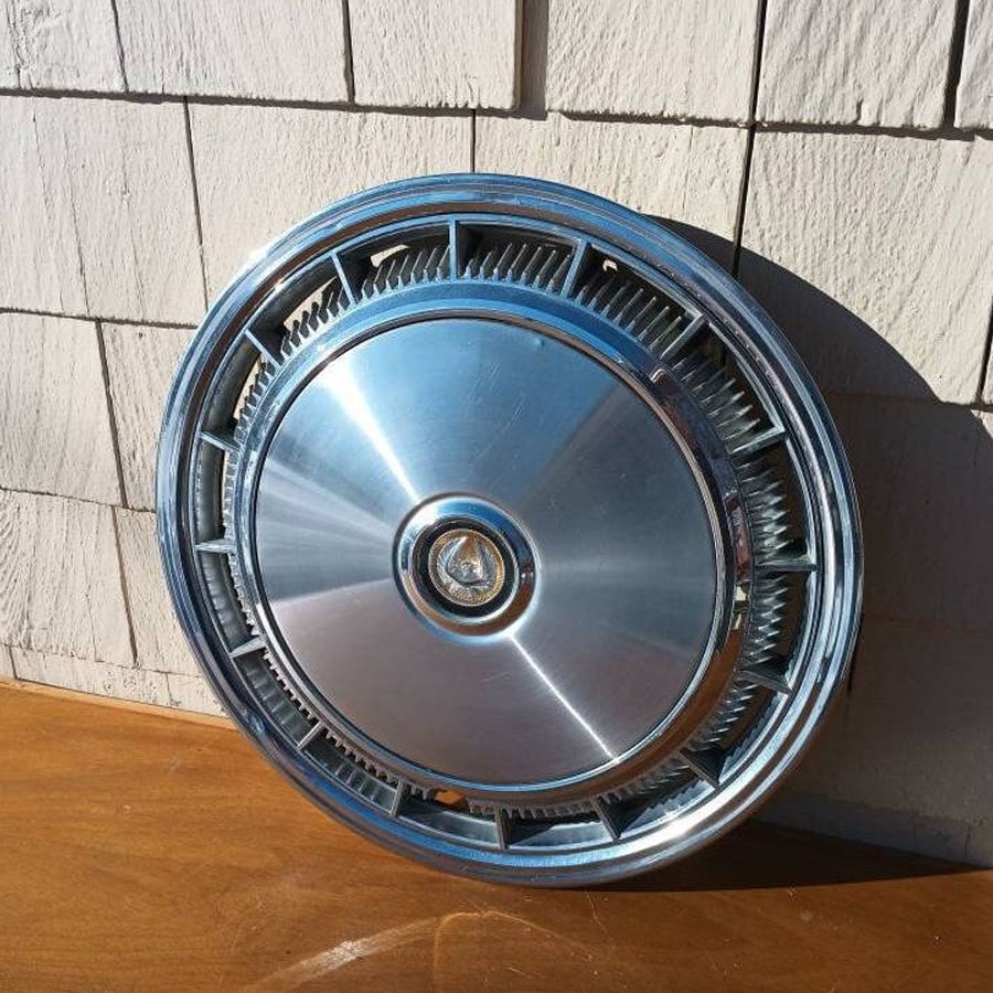 A shiny silver car hubcap with a circular design and a center emblem leans against a beige shingled wall on a wooden surface in sunlight.