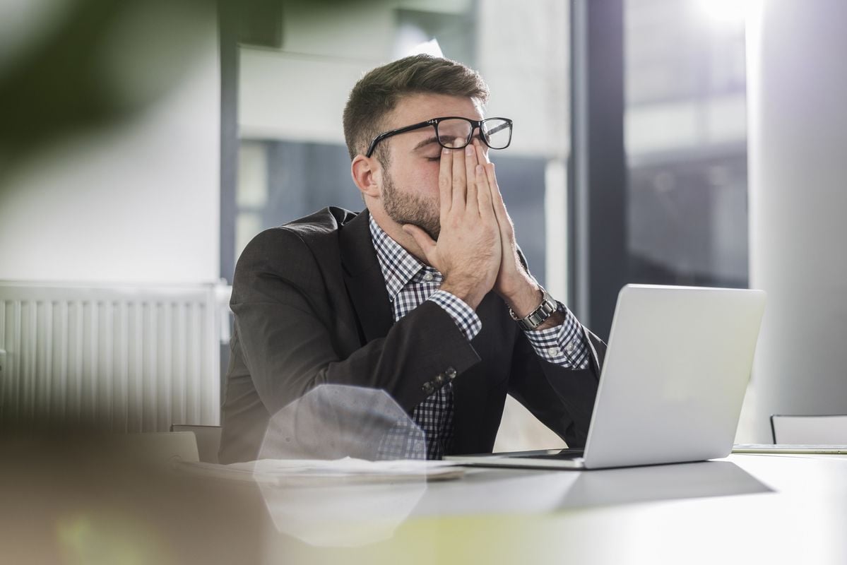 A man in a suit sits at a desk with a laptop, covering his face with both hands, appearing stressed or frustrated in a bright office setting.