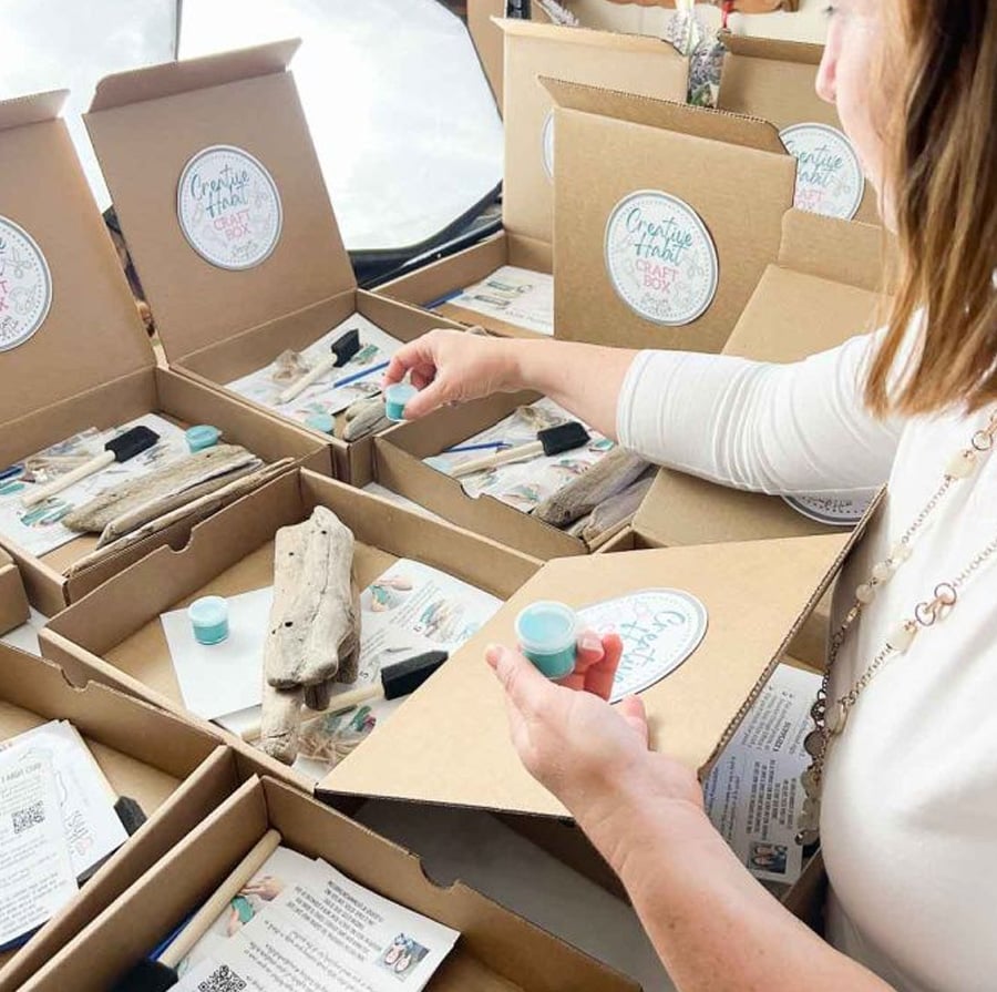 A woman assembles craft kits in open cardboard boxes, placing small containers inside. Each kit contains driftwood, a brush, and printed instructions, laid out on a table in a bright room.