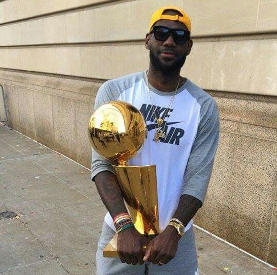 A man wearing sunglasses, a yellow cap, and a Nike Air shirt stands outside holding a large golden trophy shaped like a basketball on a pedestal.