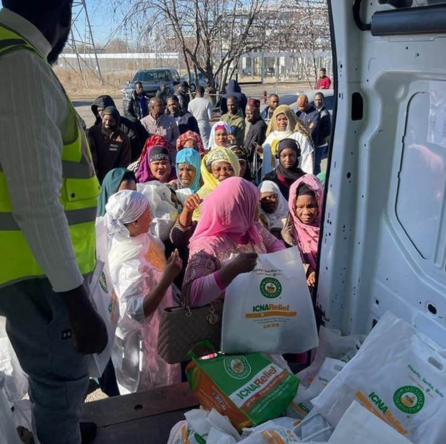 A group of people stand in line outdoors, receiving food and supply bags from volunteers at the back of a van during a community aid distribution event. The bags are labeled "ICNA Relief.