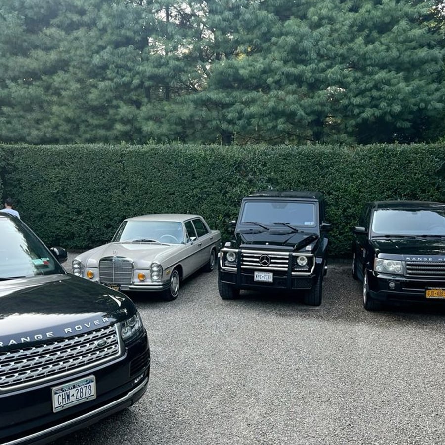 Three cars—a black Range Rover, a vintage silver Mercedes-Benz, and a black Mercedes-Benz G-Class—are parked side by side in front of green hedges and tall trees on a gravel surface.