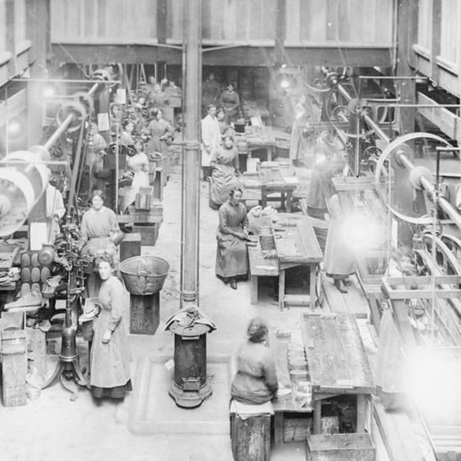 Black-and-white photo of a large factory workshop filled with women working at wooden benches and machinery, wearing long coats and aprons. Overhead pulleys, belts, and industrial equipment are visible.