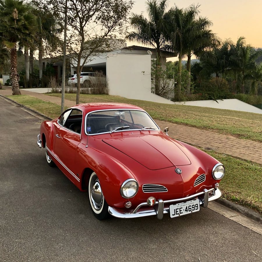 A classic red Volkswagen Karmann Ghia is parked on a suburban street near a modern house, surrounded by palm trees and greenery, under a clear evening sky.