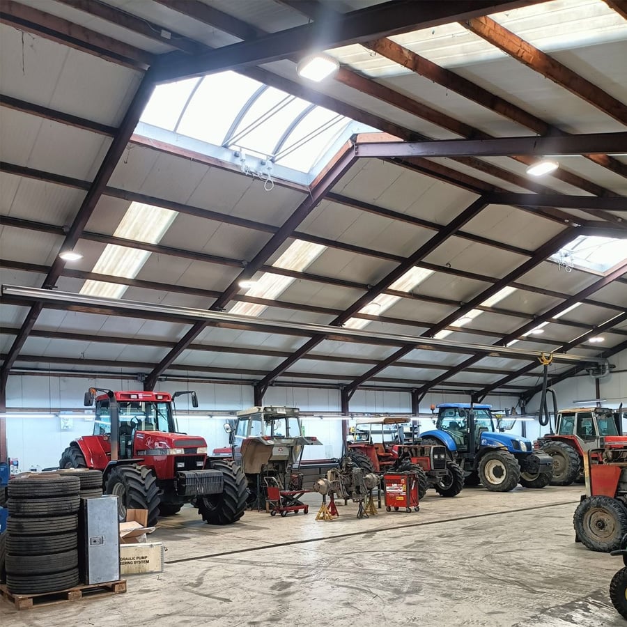 A spacious workshop with several red and blue tractors parked inside. The area is well-lit with skylights and overhead lights, and tools, equipment, and tires are scattered around the concrete floor.