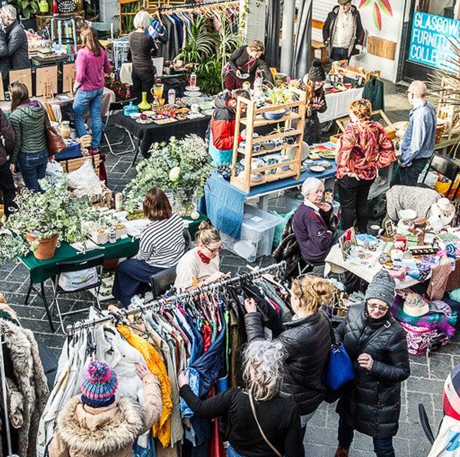 A busy indoor market scene with people browsing clothes, antiques, and crafts at various stalls. Tables are decorated with flowers and items for sale, creating a lively and colorful atmosphere.
