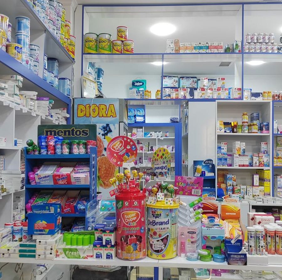 A pharmacy counter filled with various health products, medicines, and colorful candy displays, with shelves of medications and other items lining the walls in the background.