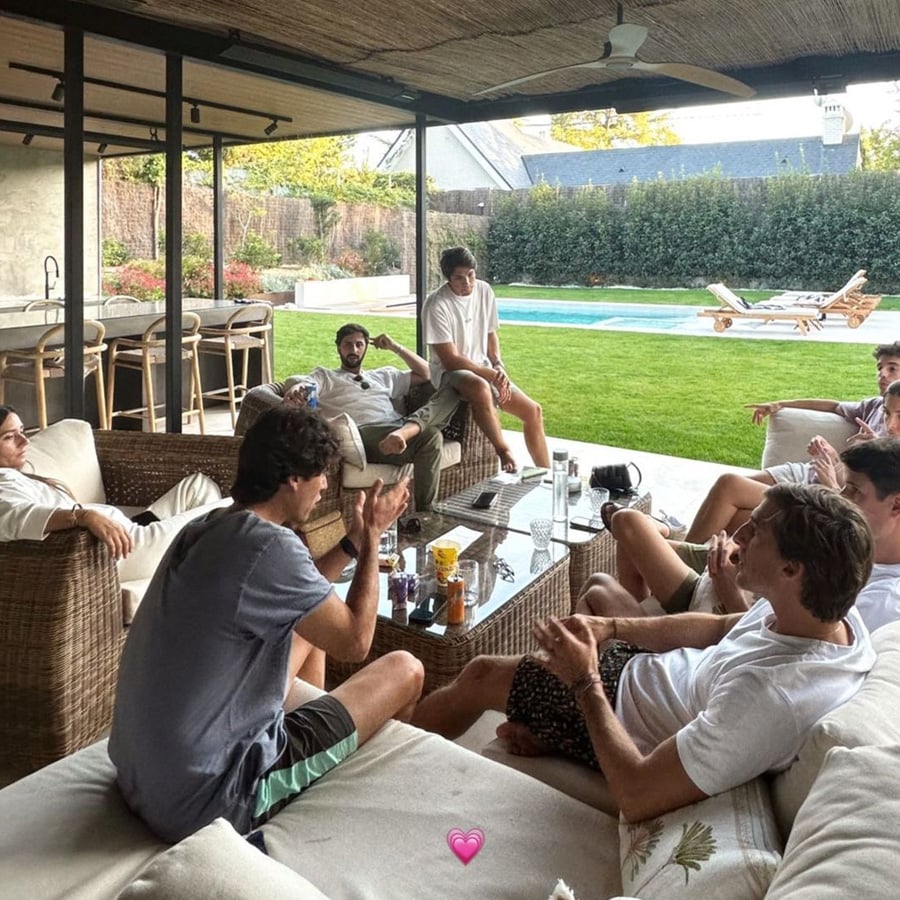 A group of people relax and chat on outdoor sofas in a covered patio area beside a swimming pool, with drinks and snacks on the table. The weather looks pleasant and the scene is casual and social.