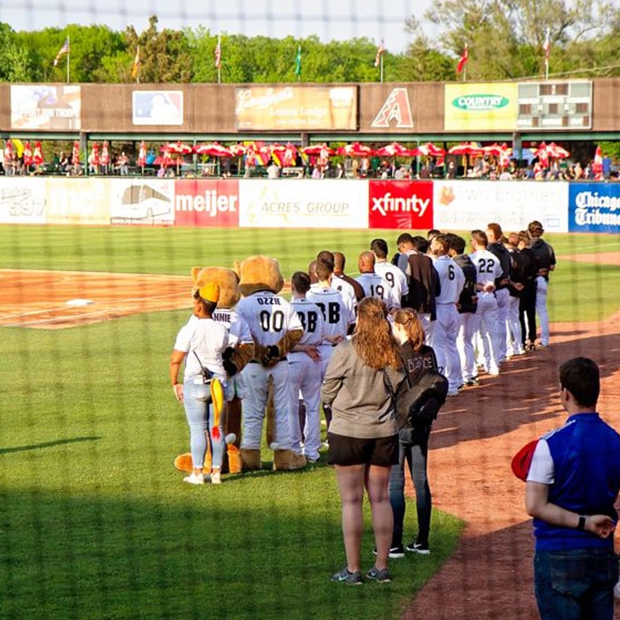 Baseball players and staff stand in a line on the field with their hands over their hearts, facing the flag for the national anthem before a game. A mascot stands among them. Spectators fill the stands in the background.