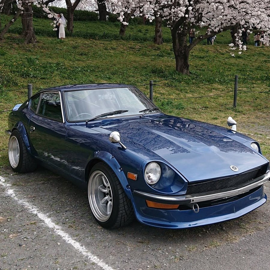 A classic blue sports car with wide tires and chrome wheels is parked on gravel near blooming cherry blossom trees and green grass.
