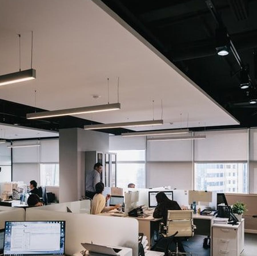 Modern open-plan office with people working at desks, computers and paperwork visible, large windows let in natural light, and ceiling lights hang above the workspace.