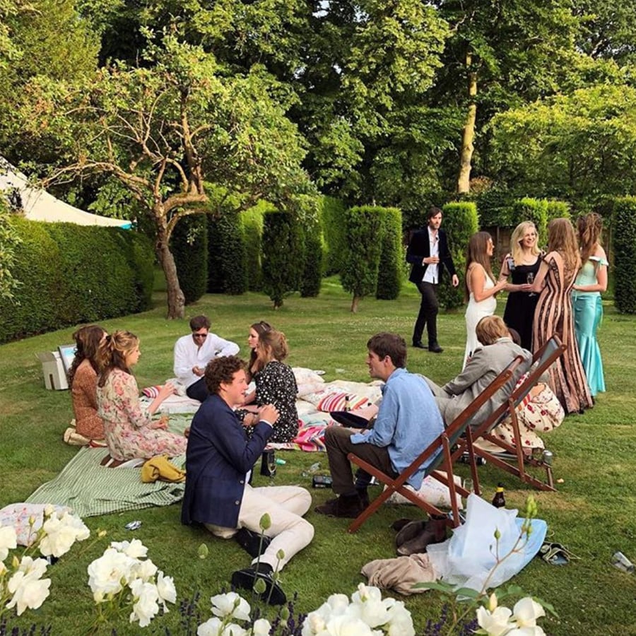 A group of people socializes outdoors on a grassy lawn, some sitting on blankets and deck chairs, others standing and talking. The scene is surrounded by trees and greenery on a sunny day.