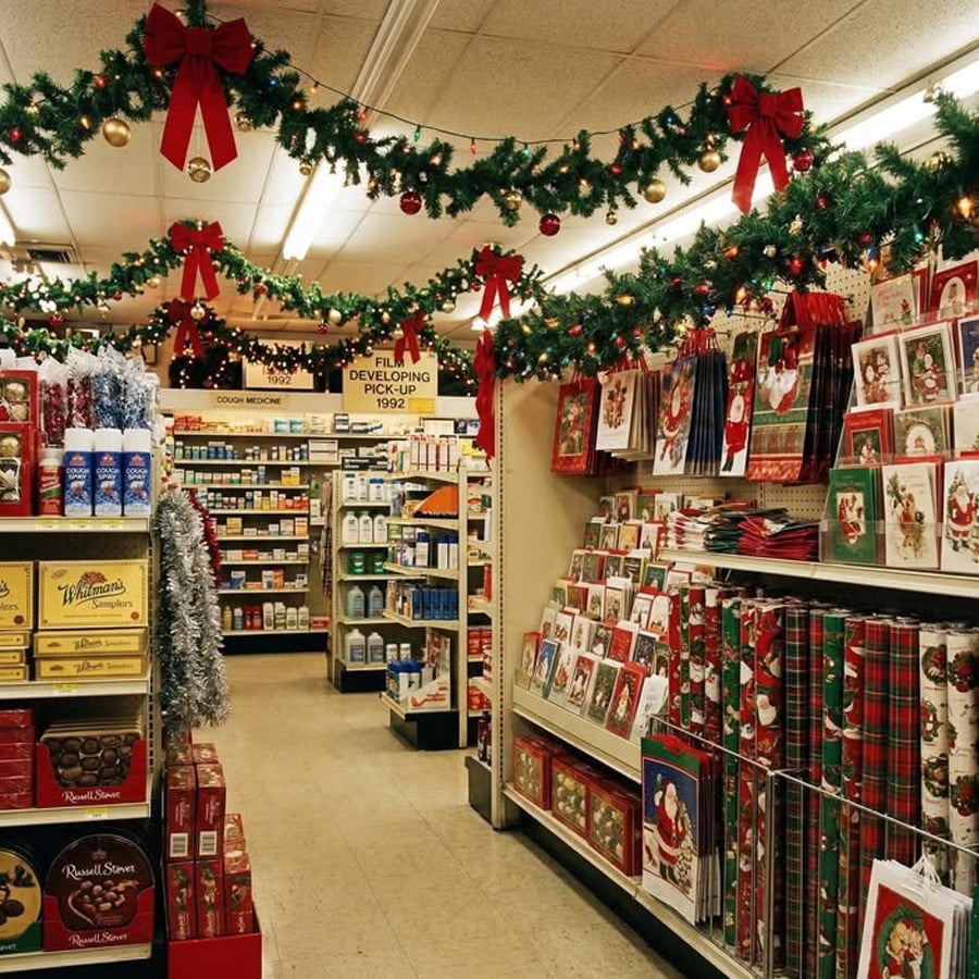 A store aisle decorated with Christmas garlands and ornaments, with shelves stocked with holiday cards, wrapping paper, chocolates, gifts, and pharmacy items. Festive decorations hang from the ceiling.
