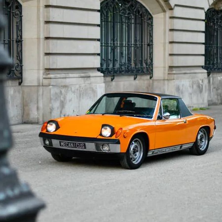 An orange vintage Porsche 914 convertible is parked on a street in front of an ornate building with large windows and wrought iron railings.