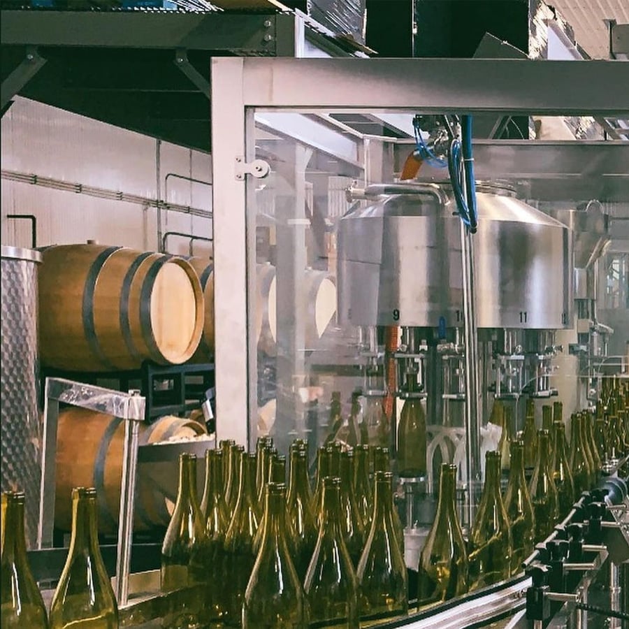 A wine bottling line shows empty glass bottles on a conveyor belt, with stainless steel machinery and wooden barrels in the background inside a winery.